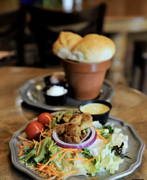 A plate of salad with tomatoes, carrots, and fried items, alongside a bread roll in a flower pot and dipping sauce.