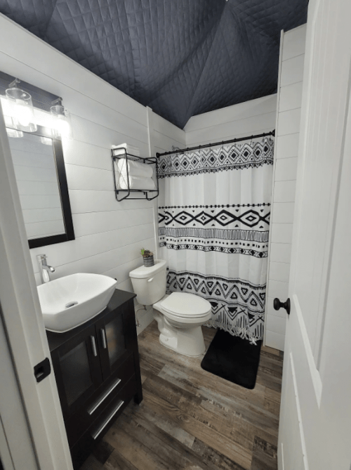 A modern bathroom featuring a black and white shower curtain, sink, toilet, and wooden flooring.