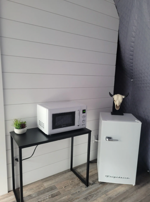 A small kitchen corner featuring a microwave, a white fridge, a black table, and a decorative skull on top.