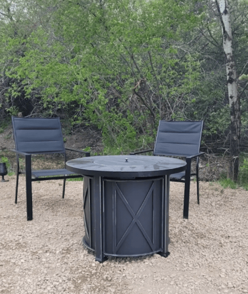 A circular fire pit surrounded by two black chairs in a natural outdoor setting with greenery.