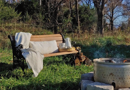 A cozy outdoor scene featuring a wooden bench with a blanket, a basket, and a fire pit surrounded by greenery.