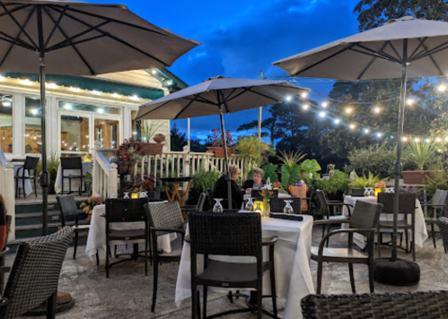 Outdoor restaurant setting with tables under umbrellas, string lights, and plants, creating a cozy evening atmosphere.