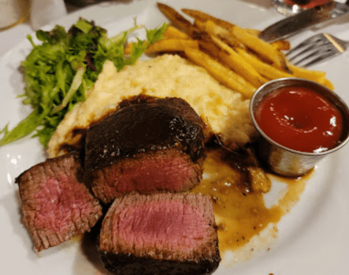 A plate featuring sliced steak, creamy mashed potatoes, a side of greens, French fries, and a small cup of ketchup.