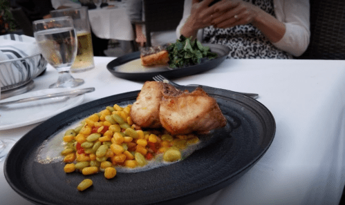 A plate of seared fish with corn and bean medley, accompanied by a glass of water and a blurred figure in the background.