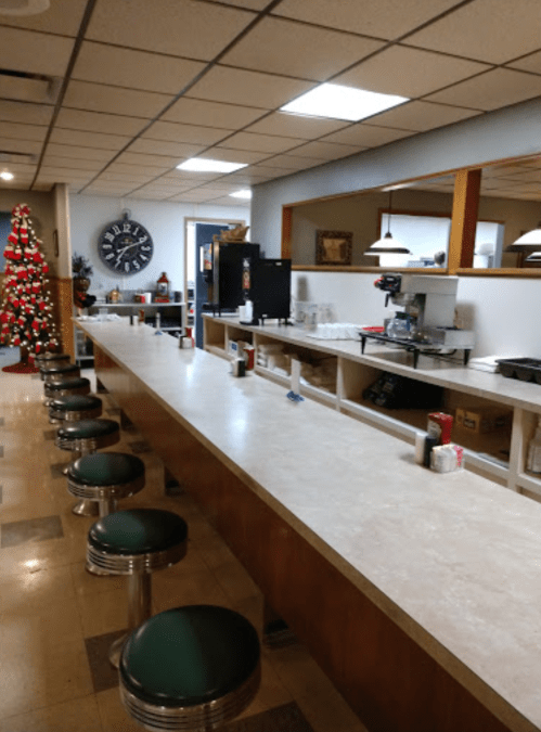 A diner interior featuring a long counter with green stools, a Christmas tree, and a clock on the wall.