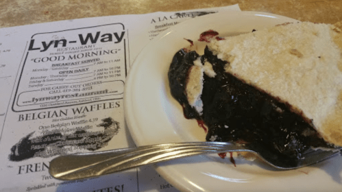 A slice of blueberry pie on a plate with a fork, next to a restaurant menu featuring breakfast options.