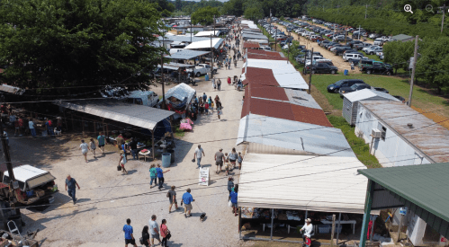 Aerial view of a bustling outdoor market with vendors, shoppers, and parked cars along a dirt path.