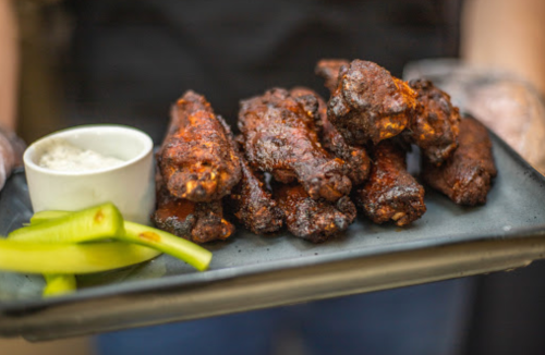 A plate of crispy, dark chicken wings served with celery sticks and a small bowl of dipping sauce.