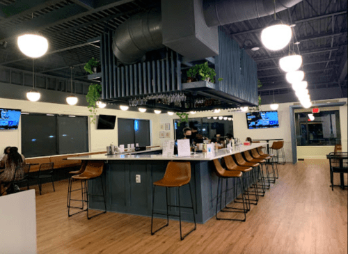 A modern bar area with high stools, hanging plants, and large windows, featuring a bartender and multiple TVs.