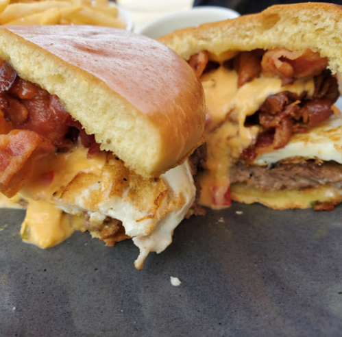 Close-up of a juicy burger with bacon, melted cheese, and a fried egg, served with fries on the side.
