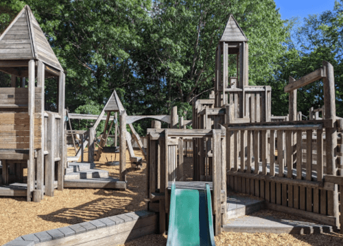 A wooden playground with slides, climbing structures, and towers, surrounded by trees and a sunny blue sky.