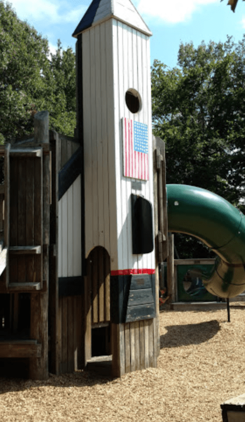 A wooden playground structure designed like a rocket, featuring a slide and an American flag.