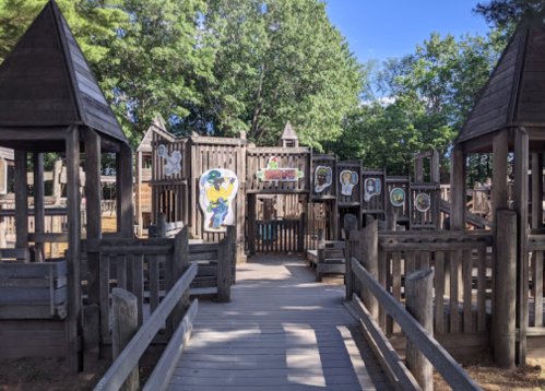A wooden playground with towers and colorful artwork on the walls, surrounded by trees and a clear blue sky.
