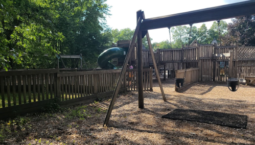 A playground with swings and a slide, surrounded by wooden fencing and trees in a sunny outdoor setting.