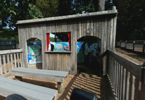 A wooden play structure with arched openings, featuring colorful murals inside and benches outside.