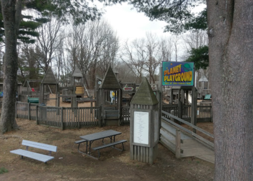 A wooden playground with various structures, benches, and a sign reading "Planet Playground" surrounded by trees.