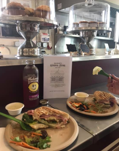 A café scene featuring two plates with sandwiches, a drink, and a menu, with cupcakes displayed in the background.