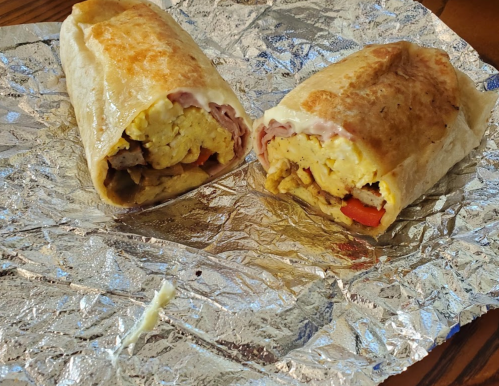 Two breakfast burritos wrapped in foil, filled with eggs, ham, and colorful peppers.