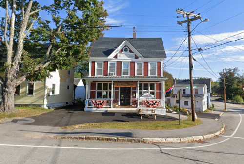 A two-story house with a porch, surrounded by trees and power lines, located at a street corner on a sunny day.
