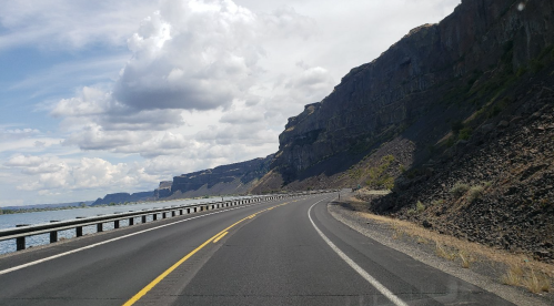 A winding road alongside a rocky cliff and water under a cloudy sky.