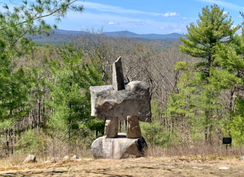 A large stone sculpture stands on a hillside, surrounded by trees and mountains in the background under a clear blue sky.