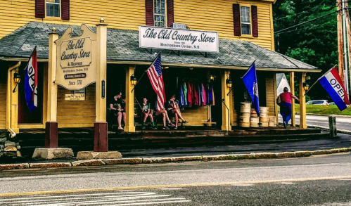 A yellow country store with an "OPEN" sign, flags, and people sitting on the porch in a rural setting.
