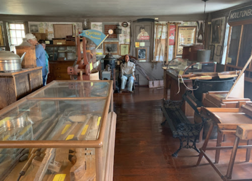 A vintage museum interior with wooden displays, artifacts, and a seated figure among historical items.