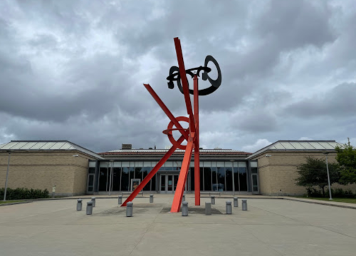 Abstract red and black sculpture with dynamic lines in front of a modern building under a cloudy sky.