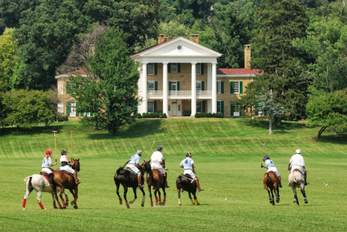A group of polo players on horseback riding across a green field in front of a large, historic mansion.