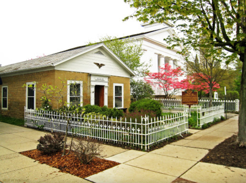 A quaint house with a white picket fence, surrounded by greenery and trees, next to a larger building.