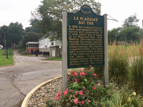 Sign for La Plaisance Bay Pike, detailing its history and significance, with a road and buildings in the background.