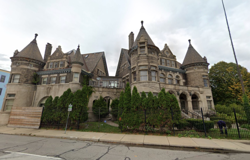 A large, ornate stone mansion with turrets and lush greenery in front, set against a cloudy sky.