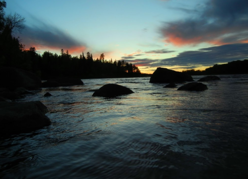 A serene river at sunset, with rocks emerging from the water and colorful clouds in the sky.