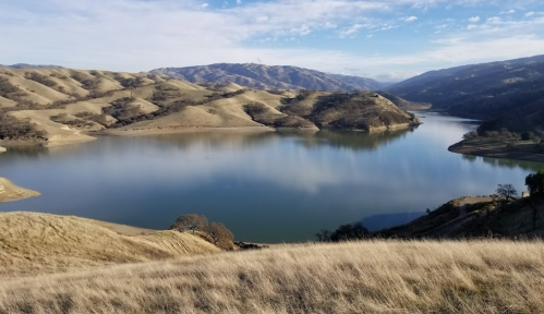 A serene landscape featuring a calm lake surrounded by rolling hills and mountains under a clear blue sky.