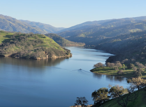 A serene river flows through green hills, surrounded by mountains under a clear blue sky.