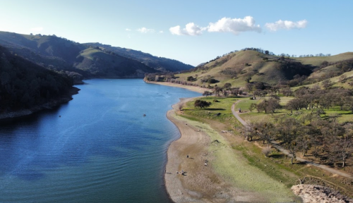 A serene landscape featuring a calm lake surrounded by rolling hills and trees under a clear blue sky.