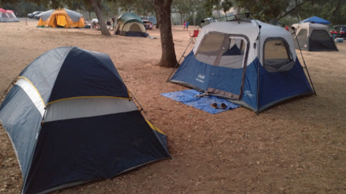 Two tents set up on a dirt campsite, surrounded by trees and other tents in the background.