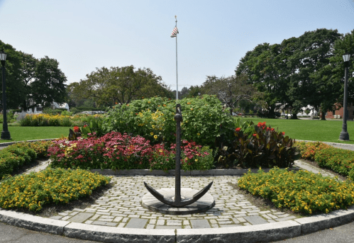 A large anchor stands in a flower garden, surrounded by colorful blooms and greenery, with a flagpole in the center.