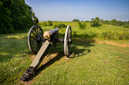 A historic cannon on a grassy field, surrounded by trees and rolling hills under a clear blue sky.