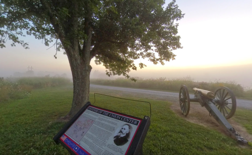 A foggy landscape featuring a historical sign and a cannon near a tree, with a road in the background.