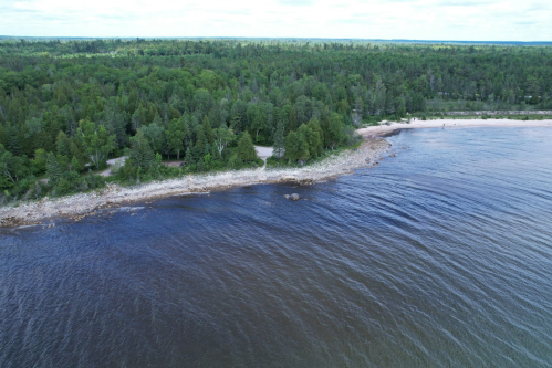Aerial view of a serene shoreline with lush green trees and calm waters, under a cloudy sky.