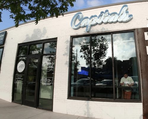 Exterior of a restaurant named "Capitals," featuring large windows and a modern sign, with a person inside.