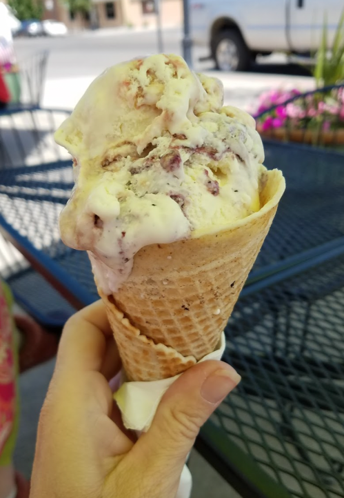 A hand holding a cone of ice cream with swirls of cream and chunks, set against a blurred outdoor background.