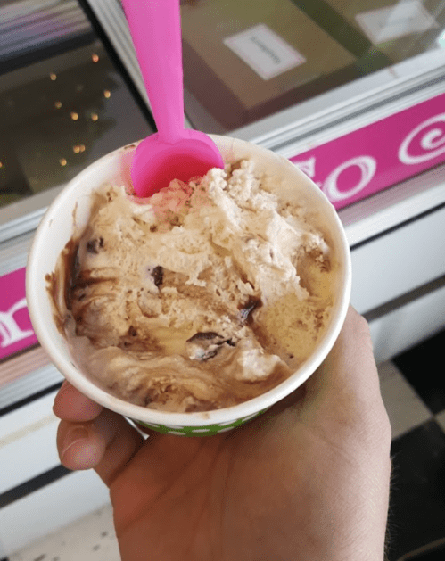 A hand holding a cup of swirled ice cream with chocolate and vanilla flavors, topped with a pink spoon.