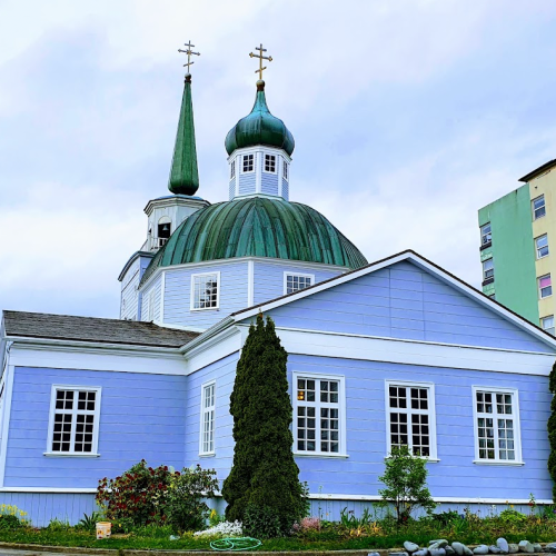 A colorful church with a green dome and spires, surrounded by greenery and a modern building in the background.