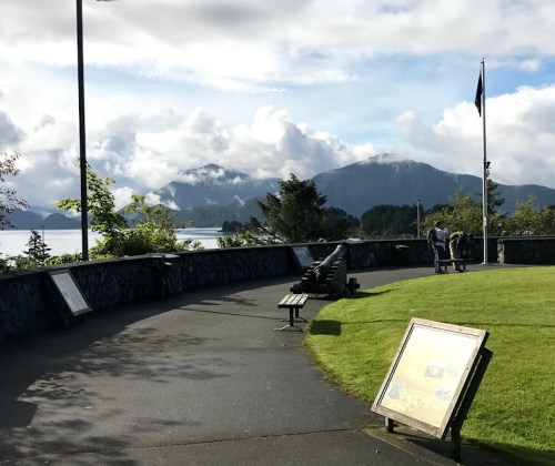 A scenic view of a pathway with a cannon, benches, and informational signs, surrounded by mountains and clouds.