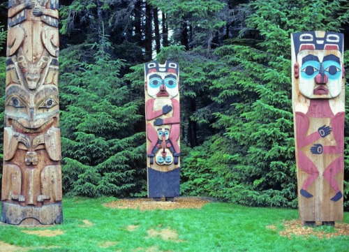 Three colorful totem poles stand among lush green trees in a natural setting.
