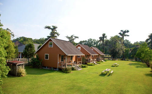 A row of cozy orange cabins on a green lawn, surrounded by trees and picnic tables under a clear blue sky.