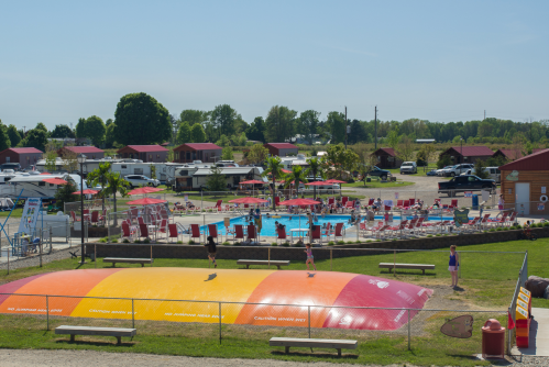 A vibrant outdoor pool area with colorful sun loungers, a trampoline, and nearby cabins under a clear blue sky.