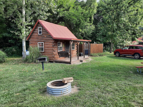 A small log cabin with a red roof, surrounded by greenery, a fire pit, and a red car parked nearby.
